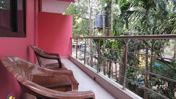 A balcony with two wooden chairs, a red wall, and a view of lush greenery and a water tank. - Pritams Cottages (Canacona)