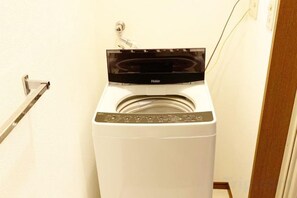 A front-loading white washer and dryer unit with a control panel on top, located in a hallway with a door on the right. - SunStay Osaki I (Tokyo)
