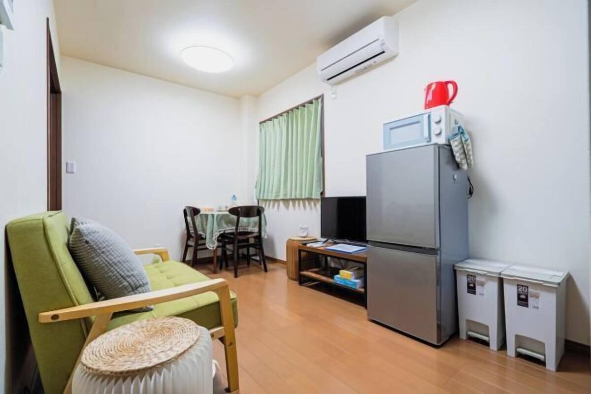 A compact living space with a sofa, a small dining table, a TV, a refrigerator, and a trash bin.