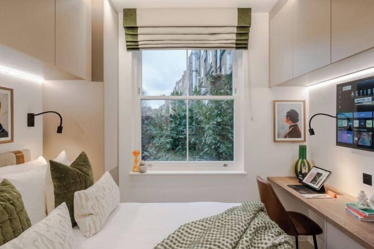 Une chambre à coucher comprenant un lit, un bureau, une chaise, une fenêtre donnant sur des arbres et un téléviseur fixé au mur.