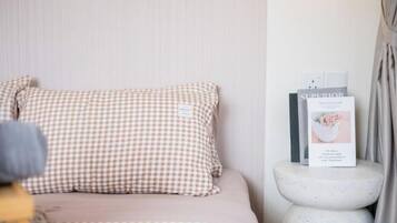Una cama con un almohadón a cuadros, un libro titulado 'SURRENDER' sobre una mesita redonda, y una pared con interruptor de luz.