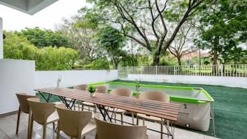 An outdoor patio with a dining table and chairs, surrounded by greenery and a swimming pool.