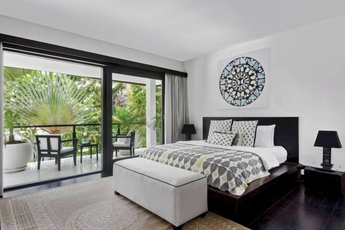 A bedroom with a large bed, a nightstand, a potted plant, and a view of greenery outside.