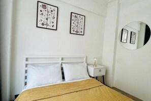 A bedroom with a white bed, two pillows, a bedside table with a lamp, and two framed floral artworks on the wall.