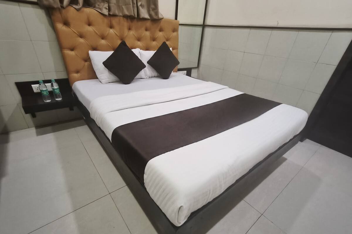 A neatly made bed with a tufted headboard, white and brown bedding, and two black pillows. There is a small bedside table with two bottles of water.