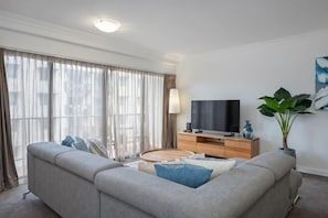 Una sala de estar moderna con un sofá, televisor, mueble de madera y ventanas amplias.