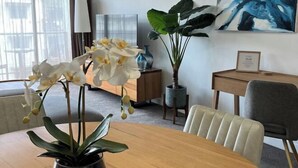 Une chambre d’hôtel avec une table à manger en bois, une orchidée en pot, une grande plante, un tableau au mur et une lampe.
