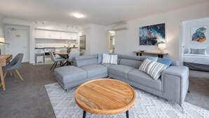 Un salon moderne avec un canapé d’angle gris, une table basse en bois et une cuisine en arrière-plan.