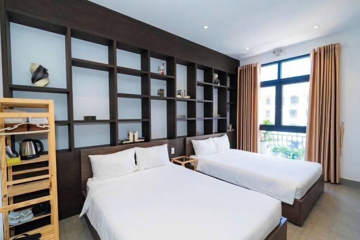 A hotel room with two beds, a wooden shoe rack, and a large window with curtains.
