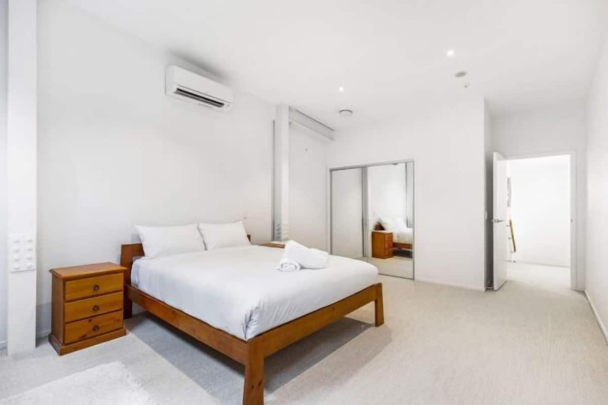 Un dormitorio con cama de madera, ropa de cama blanca, una mesita de noche, equipo de aire acondicionado, un espejo y una puerta que da a otra habitación.