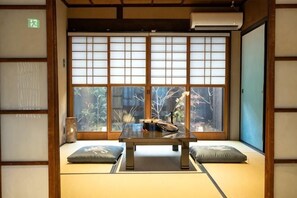 Perinteinen japanilainen huone, jossa on tatamimatto, matala pöytä, tyynyjä ja liukuovi, josta avautuu näkymä puita kohti.