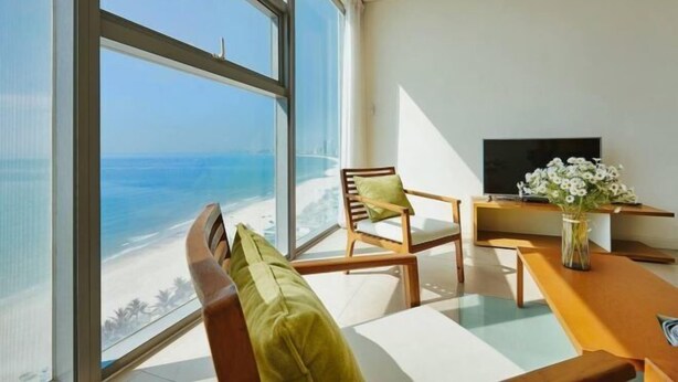 A modern room with a large window overlooking a beach, a wooden chair, a small table, and a flat-screen TV. - Beachfront 2 Bedroom Appartment with Glass wall by Villuxy (Da Nang)