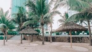 A beach area with palm trees, a thatched hut, and a stone wall. - Beachfront 2 Bedroom Appartment with Glass wall by Villuxy (Da Nang)