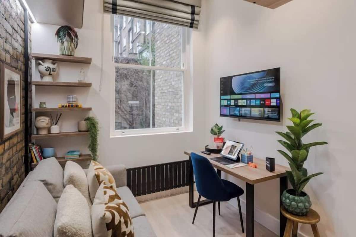Een gezellige woonkamer met een bank, een bureau met een laptop en een groot raam met jaloezieën.