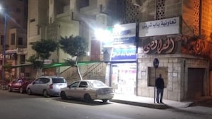 A street view with parked cars, a building with Arabic signage, and a person standing by the sidewalk. - Caesar Studio Apartment (Alexandria)