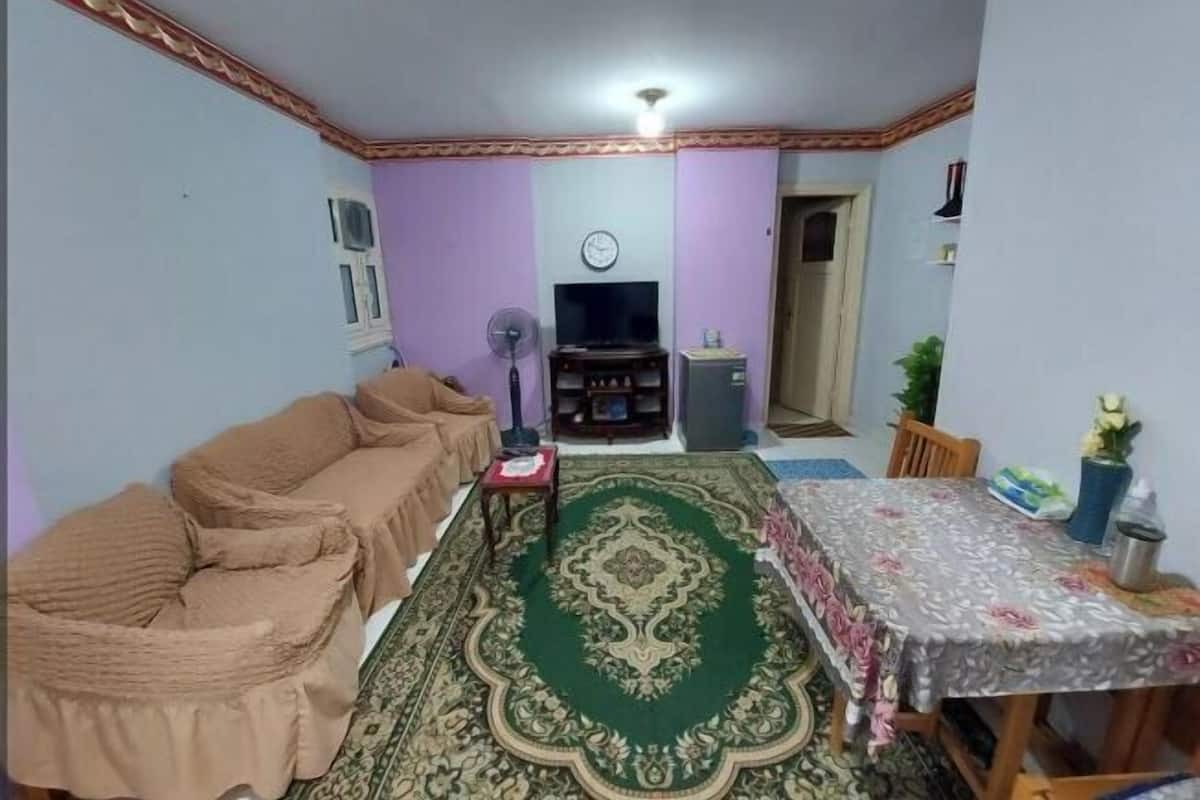 Una sala de estar con un sofá, un tapete verde estampado, un televisor y una mesa de comedor con sillas.
