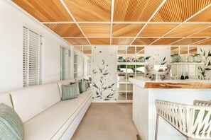 A modern interior with a wooden ceiling, a white sofa with cushions, a kitchen area with a wooden countertop, and a white chair. - All About Villa Serenity (Marbella)