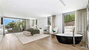 Un dormitorio amplio con una cama grande, una zona de estar con sofá y sillones, una bañera y un balcón con vistas a zonas verdes.