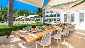 Un área de comedor al aire libre con una mesa de madera larga, sillas de mimbre blanco y sombrillas grandes.