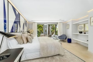 A modern bedroom with a bed, a desk, a chair, and a view of a balcony with plants.