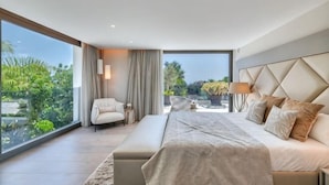 A modern bedroom with a large bed, a seating area, and a view of greenery. - All About Villa Serenity (Marbella)