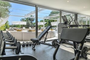 A gym with exercise equipment, a bench, and a view of a pool area with lounge chairs and umbrellas. - All About Villa Serenity (Marbella)