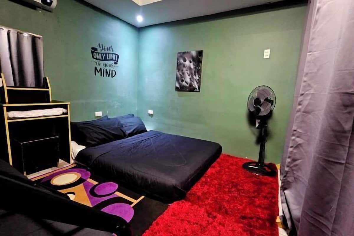 A room with a bed, a small desk, a wall with a motivational quote, a picture, and a standing fan.