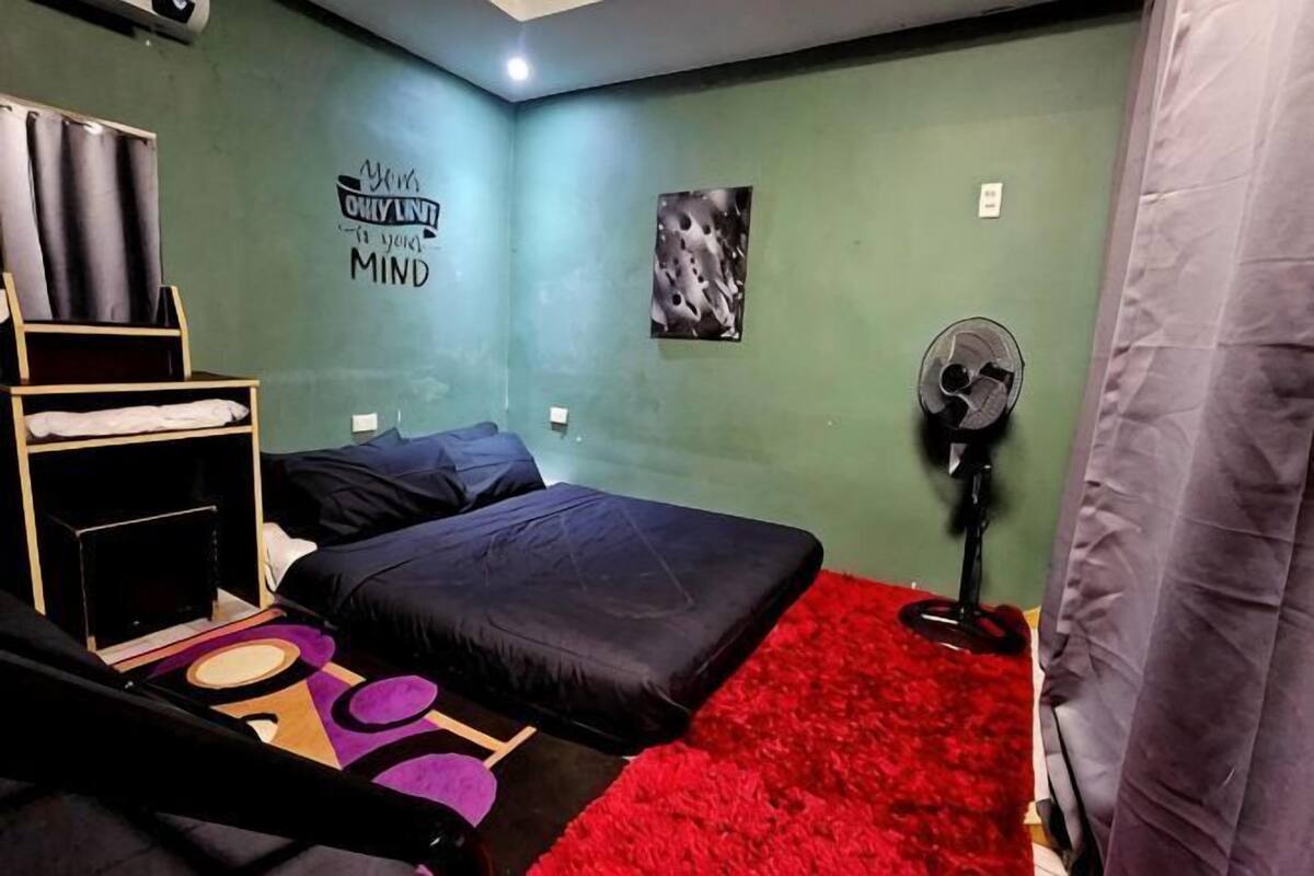 Una stanza con un letto, una scrivania, una sedia, un ventilatore e un tappeto rosso.