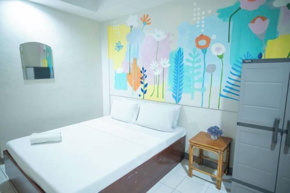 Ein Zimmer mit einem Einzelbett, einem kleinen Nachttisch, einem Spiegel und einem farbenfrohen Wandbild mit Blumen.