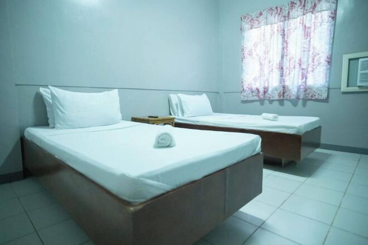 Ein Hotelzimmer mit zwei Einzelbetten, einem kleinen Nachttisch, einem Fenster mit floralem Vorhang und einer Wand-Klimaanlage.