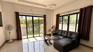 A spacious living room with a black leather sofa, a coffee table, and a floor lamp. Large windows with curtains allow natural light and views of greenery outside. - Elly's Home. Convenience & Serenity (Chiang Mai)