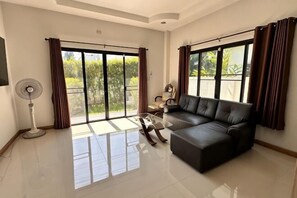 A spacious living room with a black leather sofa, a coffee table, and a floor lamp. Large windows with curtains allow natural light and views of greenery outside. - Elly's Home. Convenience & Serenity (Chiang Mai)