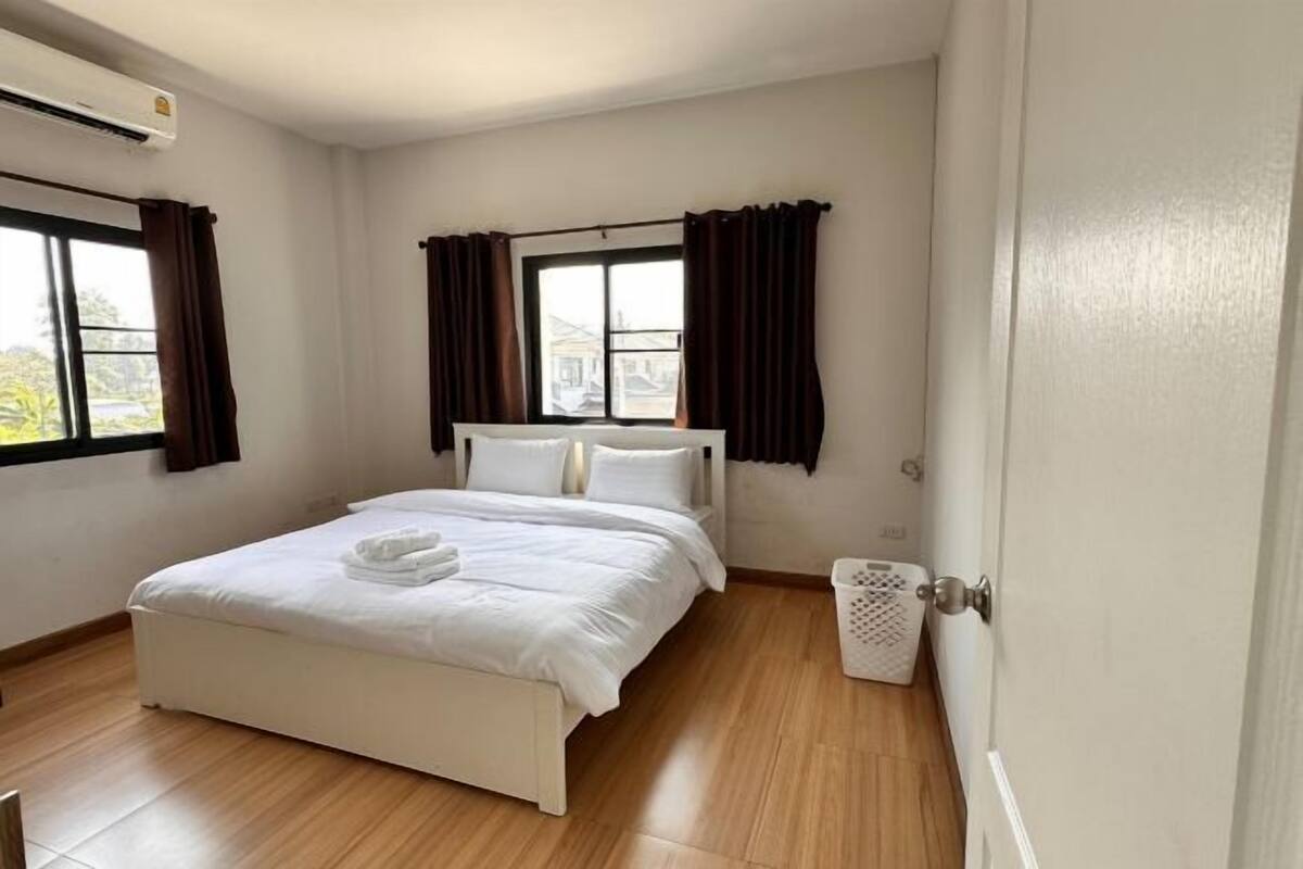 A neatly arranged bedroom with a bed, two windows with curtains, a white laundry basket, and a door.