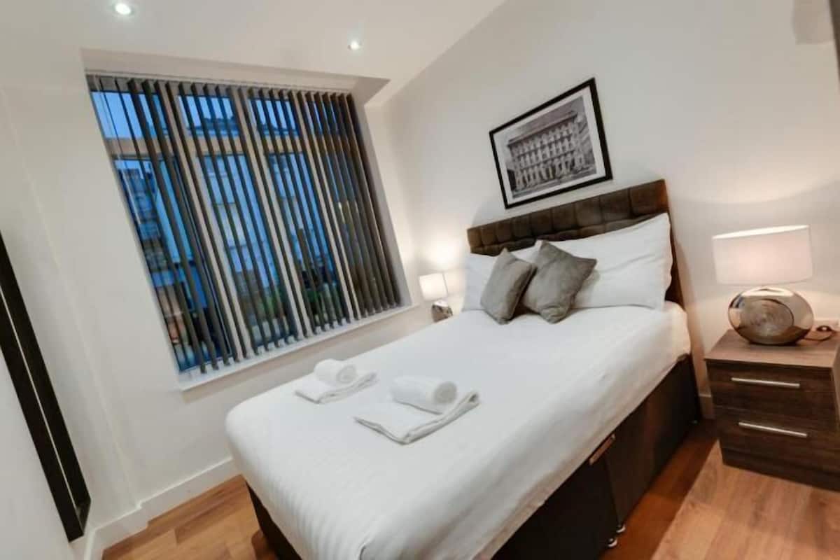 A neatly made bed with pillows and a rolled towel, a bedside table with a lamp, and a window with blinds.