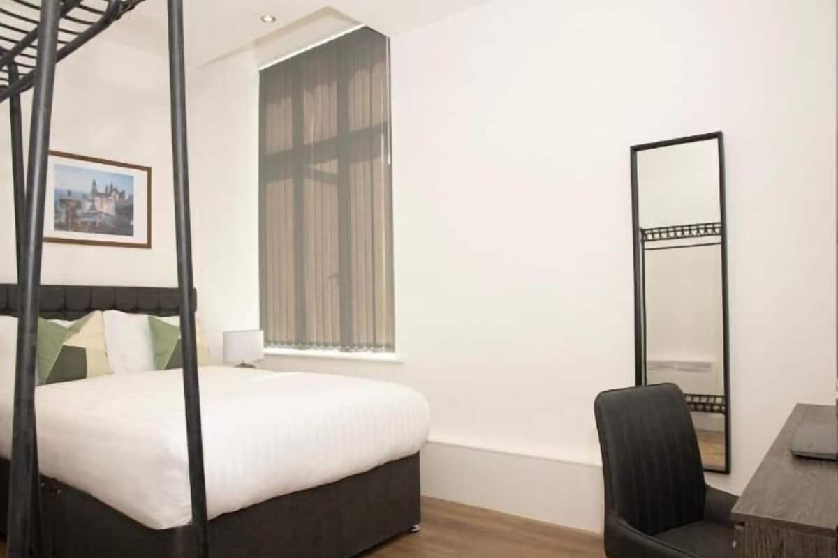A bedroom with a four-poster bed, a large mirror, a desk, and a window with blinds.