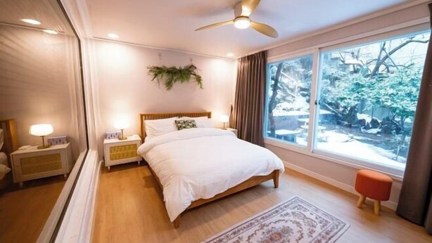 A bedroom with a bed, bedside lamps, a ceiling fan, a window with a view of trees, and a small stool. - JT_TEVA (USD) (Seoul)