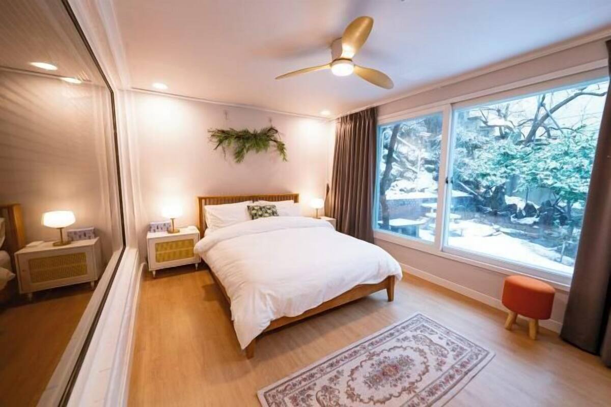 A bedroom with a bed, bedside lamps, a ceiling fan, a window with a view of trees, and a small stool.