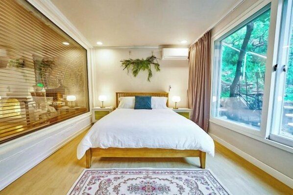 A bedroom with a large bed, bedside lamps, a window with a view of trees, and a decorative plant on the wall. - JT_TEVA (USD) (Seoul)