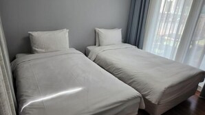 Une chambre avec deux lits simples, des murs gris et une grande fenêtre ornée de rideaux légers.