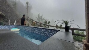 Un homme se tient près d’une piscine, adossée à un mur de pierre et bordée d’une rambarde en bois.