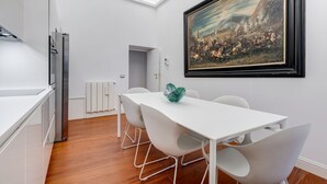 A modern kitchen with white cabinetry, a stainless steel refrigerator, and a dining area with a white table and chairs. A framed painting is on the wall above the table. - Babuino 55 - FromHometoRome (Roma)