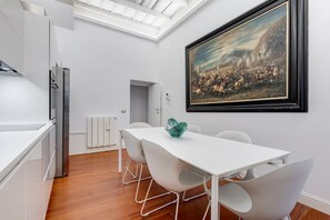 A modern kitchen with white cabinetry, a stainless steel refrigerator, and a dining area with a white table and chairs. A framed painting is on the wall above the table.