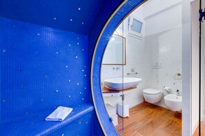 A modern bathroom with a blue tiled wall, a round mirror, a white sink, and a toilet.