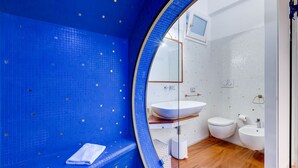 A modern bathroom with a blue tiled wall, a round mirror, a white sink, and a toilet. - Babuino 55 - FromHometoRome (Roma)