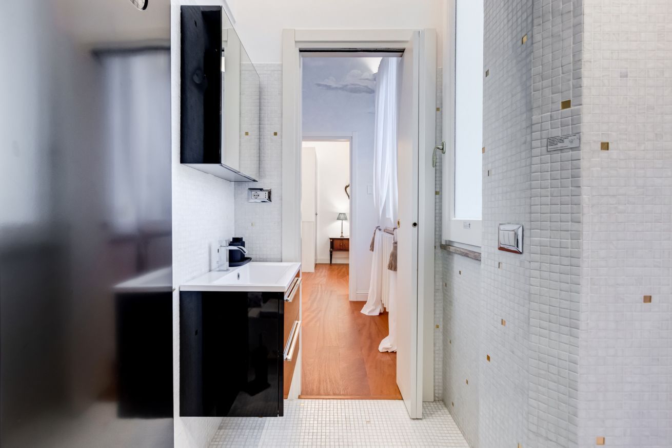 A narrow hallway with a bathroom on the left, featuring a sink and mirror. A door leads to a room with a bed and a window.