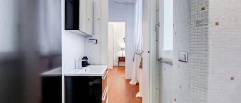A narrow hallway with a bathroom on the left, featuring a sink and mirror. A door leads to a room with a bed and a window.