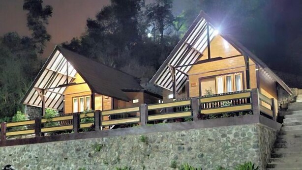 Dos cabañas modernas y bien iluminadas con techos inclinados, rodeadas de muros de piedra y barandillas de madera, con árboles al fondo.