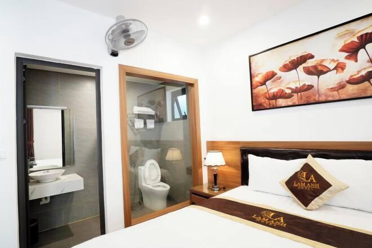 A hotel room with a bed, a bedside table, a lamp, a painting on the wall, and a bathroom with a toilet and sink.