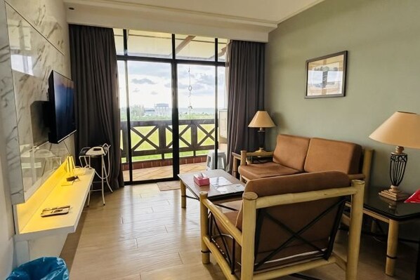 A hotel room with a sofa, a desk, a TV, a lamp, and a view through a glass door. - Heart of City Seaside view Mahkota Costa (Melaka)