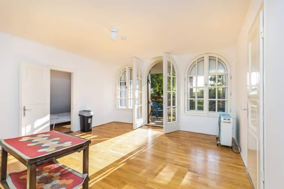 Ein Zimmer mit Holzboden, weißen Wänden und Bogenfenstern. Es gibt einen Tisch mit einer Decke, einen Mülleimer und einen Wagen.